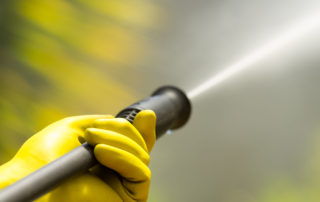 Yellow gloved hand holding black head of a high pressure water cleaner with water emerging.